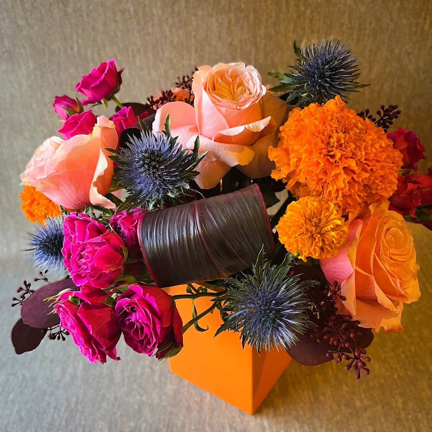 unique flower arrangement in an orange cube vase with golden marigolds, hot pink roses and purple thistle.